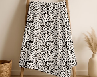 Puede incluir: Una manta blanca con estampado de leopardo, colgada de una escalera de madera. La manta presenta manchas negras y beige. Una cesta de mimbre y un jarrón con plantas secas están en el fondo.