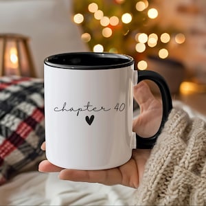 May include: White ceramic mug with a black interior and handle. The mug features the text "chapter 40" in a script font, with a small black heart below. The mug is held in a person's hand.