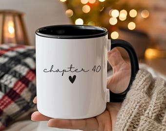 Puede incluir: Taza de cerámica blanca con interior y asa negros. La taza presenta el texto "chapter 40" en fuente cursiva, con un pequeño corazón negro debajo. La taza está sostenida en la mano de una persona.