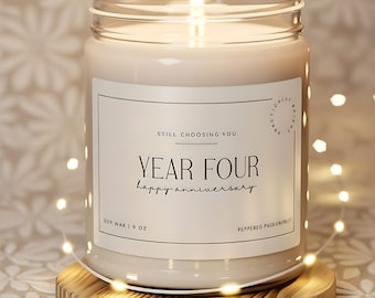May include: A lit soy wax candle in a clear glass jar, with a wooden coaster. The candle has a white label with the text "YEAR FOUR happy anniversary". The candle is 9 oz.