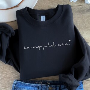 May include: Black crewneck sweatshirt with the phrase "in my phd era" in white script, with a small heart. The sweatshirt is paired with blue jeans. The Gildan brand tag is visible.