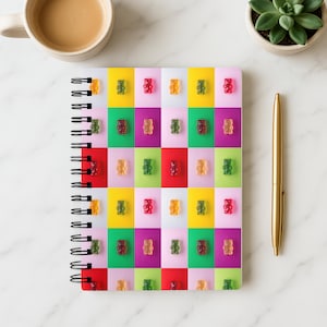 Op de afbeelding: Spiraalgebonden notitieboek met een kleurrijk rasterpatroon van gummyberen op een wit marmeren oppervlak. Het raster bevat gele, roze, groene en rode vierkanten, elk met een gummybeer. Een gouden pen, een kopje koffie en een kleine plant zijn ook aanwezig.