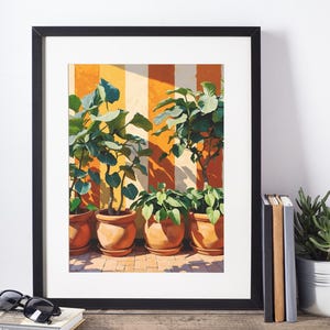 Puede incluir: Impresión artística enmarcada con plantas en macetas en macetas de terracota contra una pared a rayas naranja y crema. Las plantas tienen hojas verdes grandes. La obra de arte se exhibe sobre una superficie de madera con libros y gafas de sol.