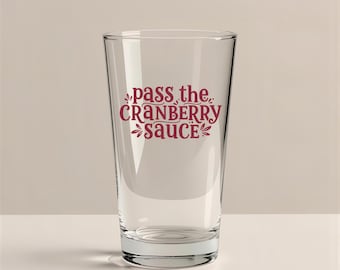 Puede incluir: Un vaso de pinta transparente con la frase "pass the CRANBERRY SAUCE" en escritura burdeos. El vaso está vacío y se encuentra sobre una superficie blanca, con un fondo neutro.