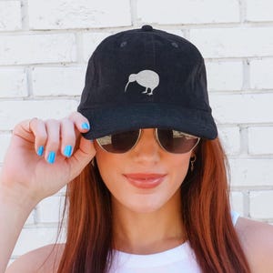 May include: Black baseball cap with a white kiwi bird design. The cap has a curved brim and a button on top. The kiwi bird is a flightless bird native to New Zealand. The cap is made of a soft, textured fabric.
