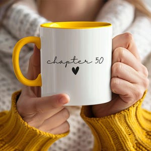 Peut inclure: Mug en céramique blanche avec intérieur et anse jaunes. Le mug porte l'inscription "chapter 50" en écriture cursive, avec un cœur noir en dessous. Le mug est tenu par une personne portant un pull jaune.