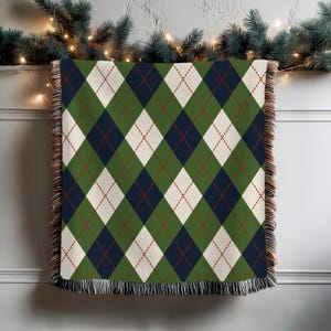 Könnte beinhalten: Eine grüne, marineblaue, cremefarbene und rote Argyle-Muster-Wurfdecke. Die Decke hat einen Fransenrand und wird vor einem weißen Hintergrund mit Weihnachtsgrün und Lichtern präsentiert.