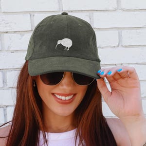 May include: Olive green corduroy baseball cap featuring a white kiwi bird design. The cap has a curved brim and a fabric strap closure. The kiwi bird is embroidered on the front of the cap.