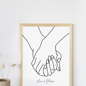 Pode incluir: Uma impressão emoldurada com um desenho minimalista de duas mãos dadas. A obra de arte é em preto e branco, com os nomes "Ana & Mário" e a data "25.04.2018" abaixo. A moldura é de madeira clara.