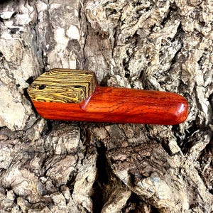 Puede incluir: Una pipa de madera hecha a mano con un cuenco marrón rojizo y un bloque con un patrón amarillo y negro contrastante para la boquilla. La pipa está colocada sobre un fondo de corteza marrón texturizada.