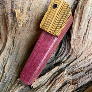 May include: A handcrafted wooden pipe with a vibrant purple body and a contrasting yellow and black striped top. The pipe features a smooth, polished finish and a small hole on the top. The background is a textured piece of wood.