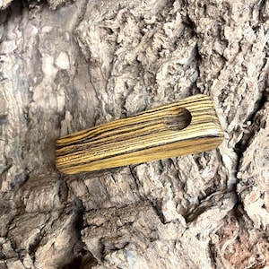May include: A rectangular wooden pipe with a circular hole. The pipe is made of wood with a zebra-like pattern of dark brown and light brown stripes. The pipe is laying on a textured, brown surface.