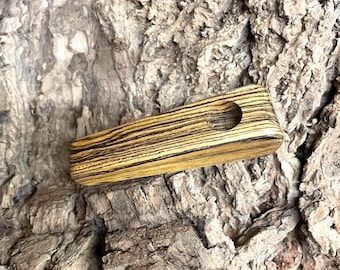 May include: A rectangular wooden pipe with a circular hole. The pipe is made of wood with a zebra-like pattern of dark brown and light brown stripes. The pipe is laying on a textured, brown surface.