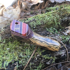 May include: A wooden slingshot with a multi-colored wood grain pattern. The slingshot has a unique design with a small hole at the top. The slingshot is resting on a mossy log.