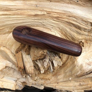 May include: A smooth, dark brown wooden pipe with a polished finish and a rounded, cylindrical body. The pipe features a circular bowl indentation near one end, showcasing a deep, rich wood grain pattern throughout the entire length of the object.