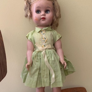May include: A vintage doll with blonde hair, blue eyes, and a green dress with a yellow ribbon. The doll is wearing blue shoes and is standing on a wooden surface.