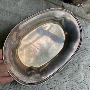May include: An oval-shaped silver-toned serving dish with a smooth, reflective surface and a slightly raised, rounded rim. The metal tray features a polished finish with subtle signs of natural oxidation and a simple, clean, unadorned design.