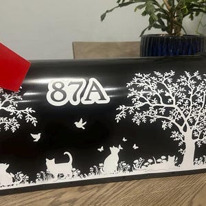 May include: Black mailbox with a white tree and cat design. The mailbox has a red flag and the number 87A on the front.
