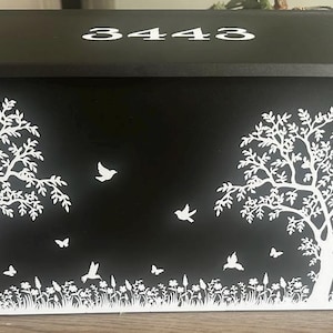 May include: A black rectangular mailbox featuring a white graphic print of two trees, flying birds, butterflies, and a field of flowers. The number 3443 is printed in white on the top lid of the mailbox, which has a wide, flat, and angled design.