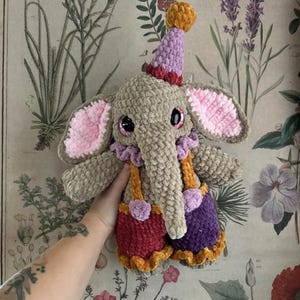 May include: A crocheted elephant doll wearing a circus outfit. The elephant is tan with pink-lined ears, large black eyes, and a purple and orange party hat. It wears red and purple shorts with orange braces.
