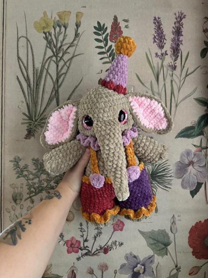 May include: A crocheted elephant doll wearing a circus outfit. The elephant is tan with pink-lined ears, large black eyes, and a purple and orange party hat. It wears red and purple shorts with orange braces.
