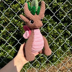 May include: A brown and pink crocheted dragon with green leaves on its head. The dragon is being held in a hand.