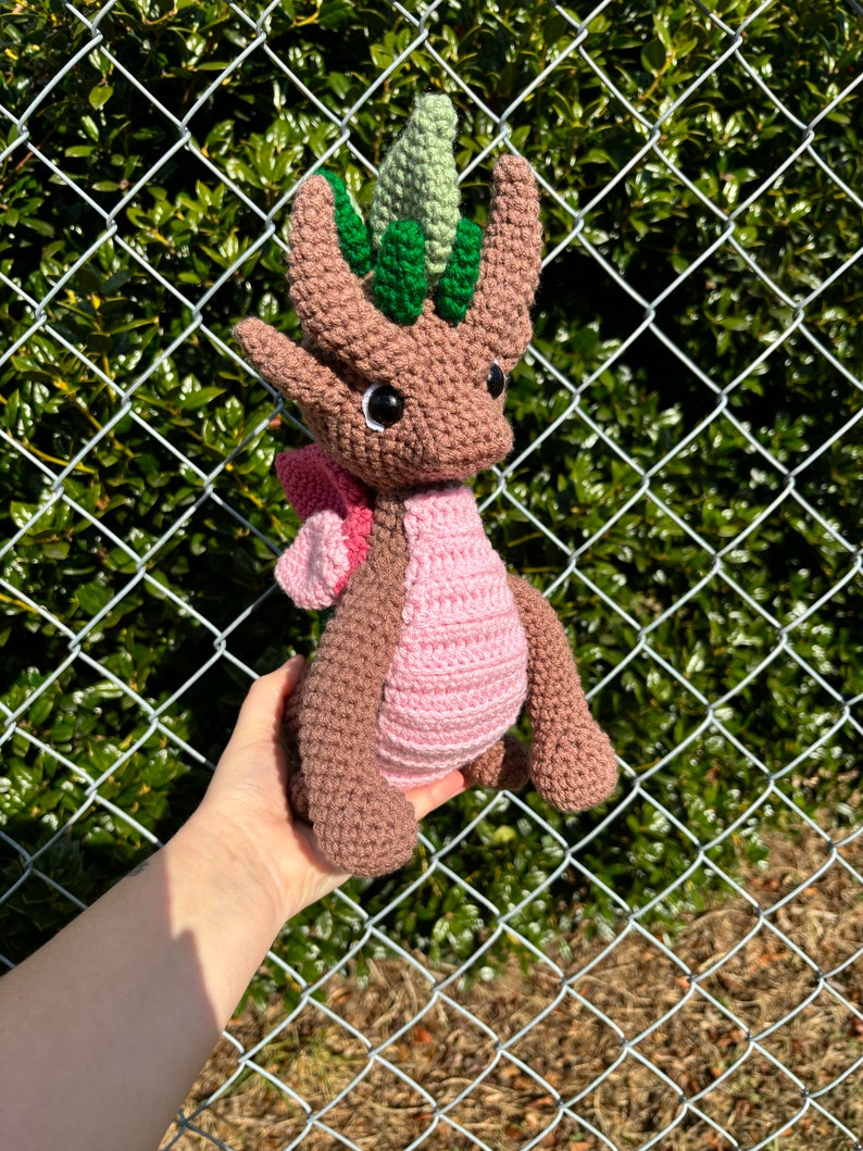 May include: A brown and pink crocheted dragon with green leaves on its head. The dragon is being held in a hand.