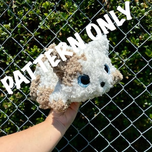 Pode incluir: Um brinquedo de pelúcia de gato branco e marrom com olhos azuis. O gato está sendo segurado na frente de uma cerca de corrente. O texto "PATTERN ONLY" está escrito em letras grandes no topo da imagem.