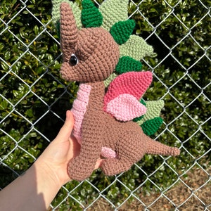May include: A brown crocheted dinosaur with green and pink spikes. The dinosaur has a black eye and a pink tongue.