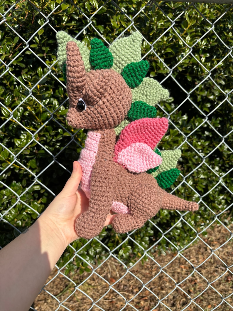 May include: A brown crocheted dinosaur with green and pink spikes. The dinosaur has a black eye and a pink tongue.