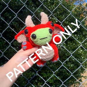 May include: A red and green crocheted plush toy with horns, wings, and two eyes. The toy is being held in a hand in front of a chain link fence.