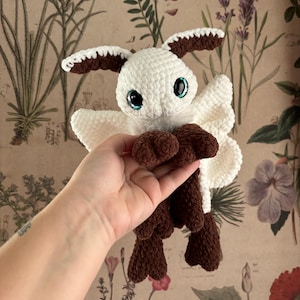 May include: A handmade crochet moth plushie. The moth has a white body, brown accents on the ears and feet, and large blue eyes. The wings are white and the plushie is held against a floral wallpaper background.