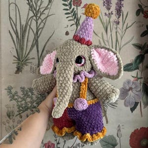 May include: A handmade crochet elephant doll wearing a party hat. The elephant is mainly grey with pink ears, large black eyes, and a long trunk. It wears purple shorts, red and orange accents, and suspenders. The doll is soft and plush.