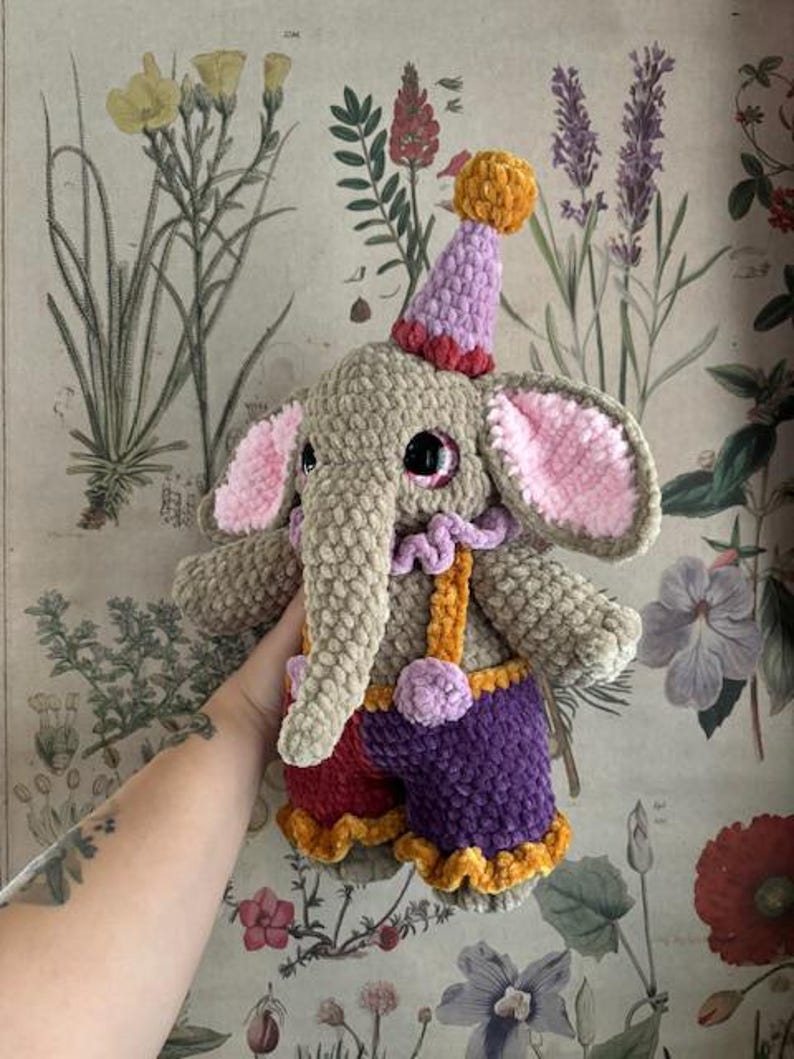 May include: A handmade crochet elephant doll wearing a party hat. The elephant is mainly grey with pink ears, large black eyes, and a long trunk. It wears purple shorts, red and orange accents, and suspenders. The doll is soft and plush.