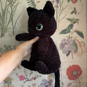 May include: A black crocheted cat plush toy with green glitter eyes. The cat is sitting on a floral patterned background.