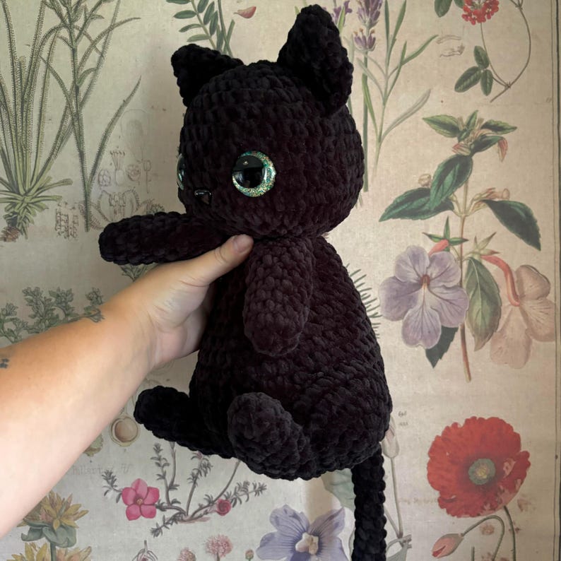 May include: A black crocheted cat plush toy with green glitter eyes. The cat is sitting on a floral patterned background.