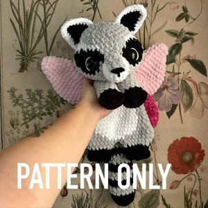 Puede incluir: Peluche de mapache tejido a ganchillo con alas rosas. El peluche tiene un cuerpo gris y blanco con detalles negros alrededor de los ojos, las orejas y las patas. El texto "PATTERN ONLY" se muestra en blanco en la parte inferior de la imagen.
