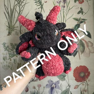 May include: A crocheted plush dragon toy with black and pink yarn. The dragon has a grumpy expression with large eyes and a long, curled tail.