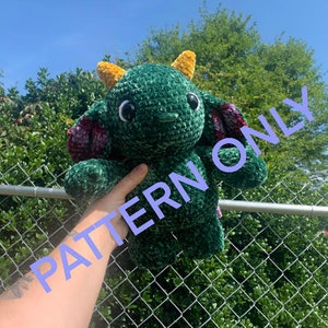May include: A green crocheted dragon plush toy with yellow horns and pink accents. The toy is being held by a hand in front of a chain link fence.
