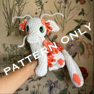 Könnte beinhalten: Ein gehäkeltes Plüschtier-Drache mit weißem und orangefarbenem Fell. Der Drache hat einen langen Schwanz und ein Paar weiße Pfeifenreiniger-Antennen. Der Drache sitzt auf einer Hand.