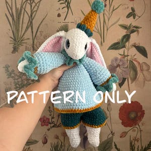 Puede incluir: Una muñeca de conejito de ganchillo hecha a mano con un diseño caprichoso. El conejito presenta una cabeza blanca, orejas forradas de rosa y un atuendo azul con detalles turquesa y naranja. El texto "PATTERN ONLY" es visible.
