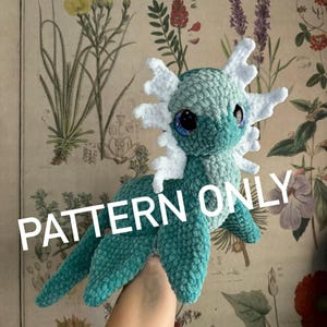 May include: A handmade teal and white crochet creature with large blue eyes. The plush toy has a textured, fuzzy appearance and features white frills around its head. The text "PATTERN ONLY" is visible on the image.