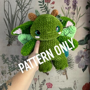 May include: A green crocheted plush toy with large, round eyes and small horns. The toy has wings with pink and blue accents. The text "PATTERN ONLY" is overlaid on the image. The background features a floral botanical print.