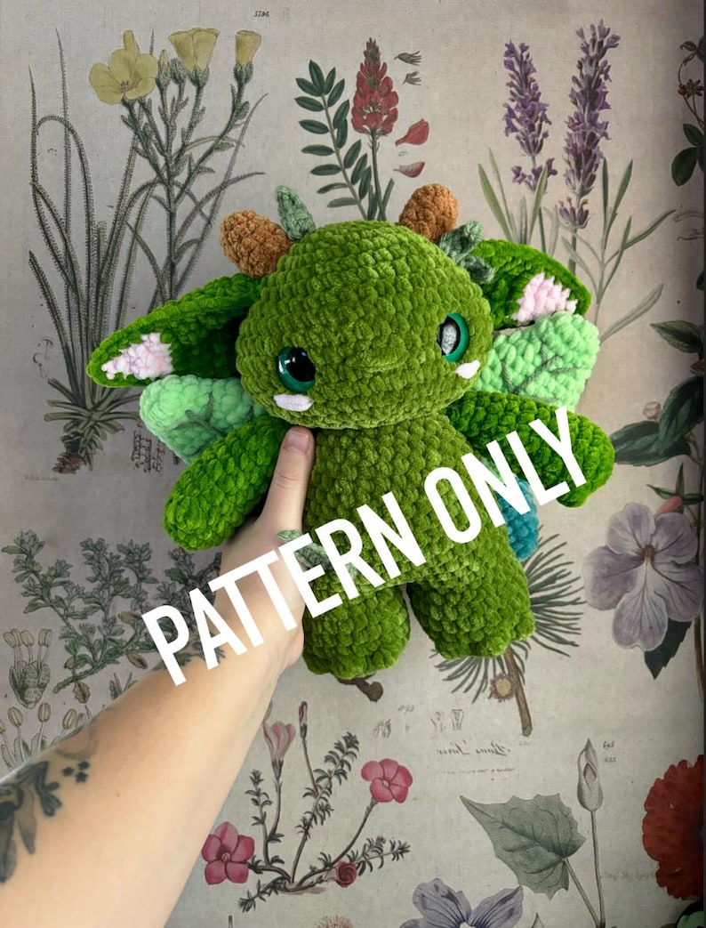 May include: A green crocheted plush toy with large, round eyes and small horns. The toy has wings with pink and blue accents. The text "PATTERN ONLY" is overlaid on the image. The background features a floral botanical print.