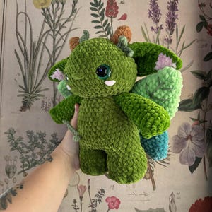 May include: A handmade green crochet dragon plushie with large, round, black eyes and small white teeth. The dragon has small orange horns, green wings with pink accents, and a small tail. The plushie is made of soft, textured yarn.