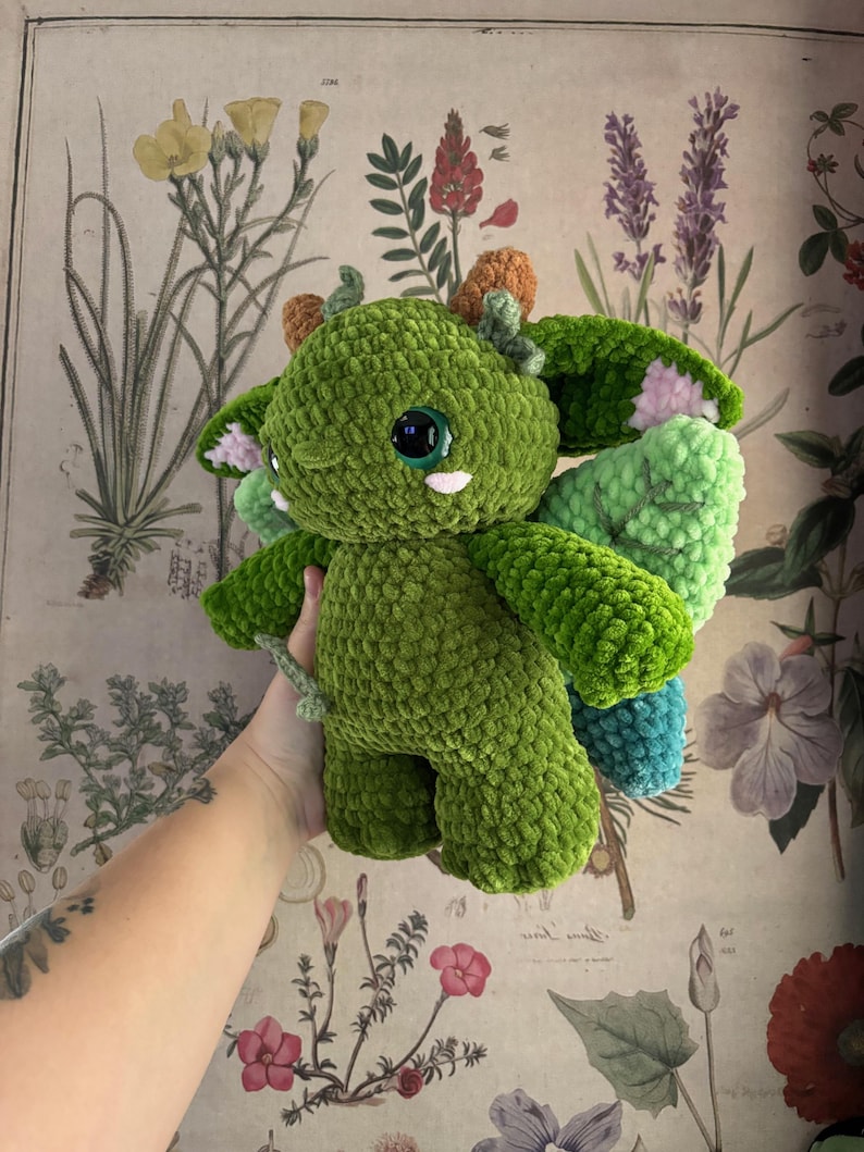 May include: A handmade green crochet dragon plushie with large, round, black eyes and small white teeth. The dragon has small orange horns, green wings with pink accents, and a small tail. The plushie is made of soft, textured yarn.