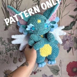May include: A teal crocheted plush creature with large pointed ears, white wings, and a yellow belly patch. The plush features blue plastic eyes, a small yellow star on its forehead, and the text PATTERN ONLY printed across the top.