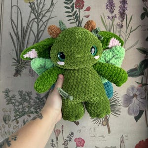 May include: A handmade green crochet dragon plushie with large, round eyes and small wings. The dragon has accents of pink, orange, and blue. The plushie is held in a person's hand against a floral background.