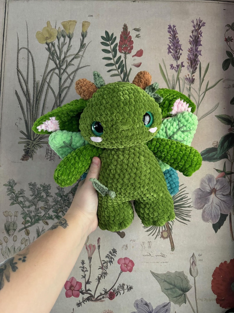 May include: A handmade green crochet dragon plushie with large, round eyes and small wings. The dragon has accents of pink, orange, and blue. The plushie is held in a person's hand against a floral background.