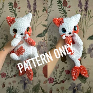 Puede incluir: Un peluche de gato hecho a mano en crochet blanco y naranja. El gato tiene ojos grandes y expresivos y una cola de pez. El texto "PATTERN ONLY" está impreso en la imagen. El peluche se sostiene sobre un fondo floral.